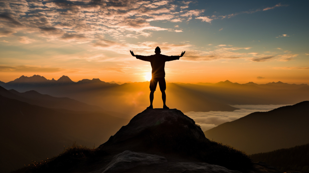 Um homem no topo da montanha de frente para um pôr do sol. A imagem representa propósito como motivação.