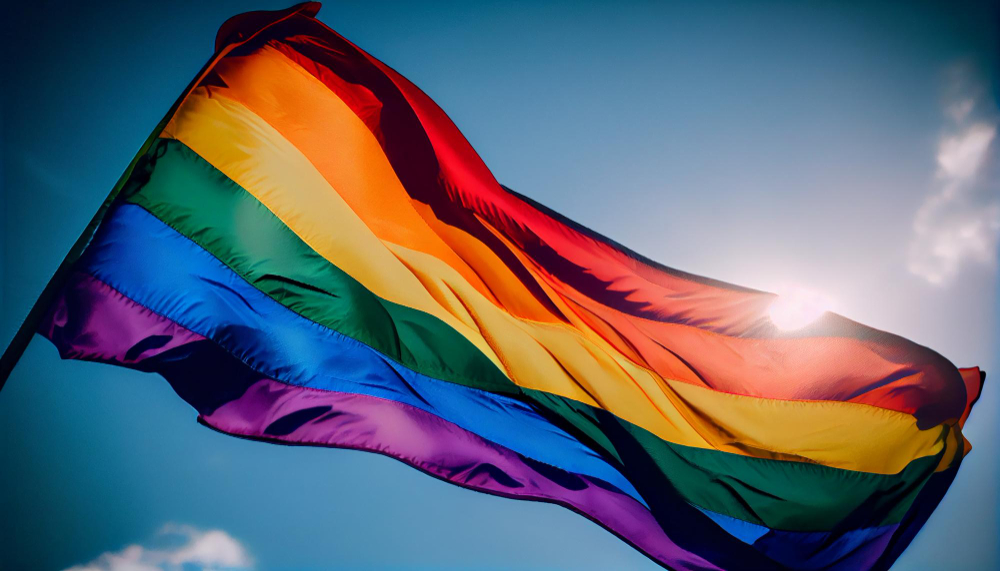A bandeira LGBTQIAPN+ do orgulho, e no fundo, um céu ensolarado.